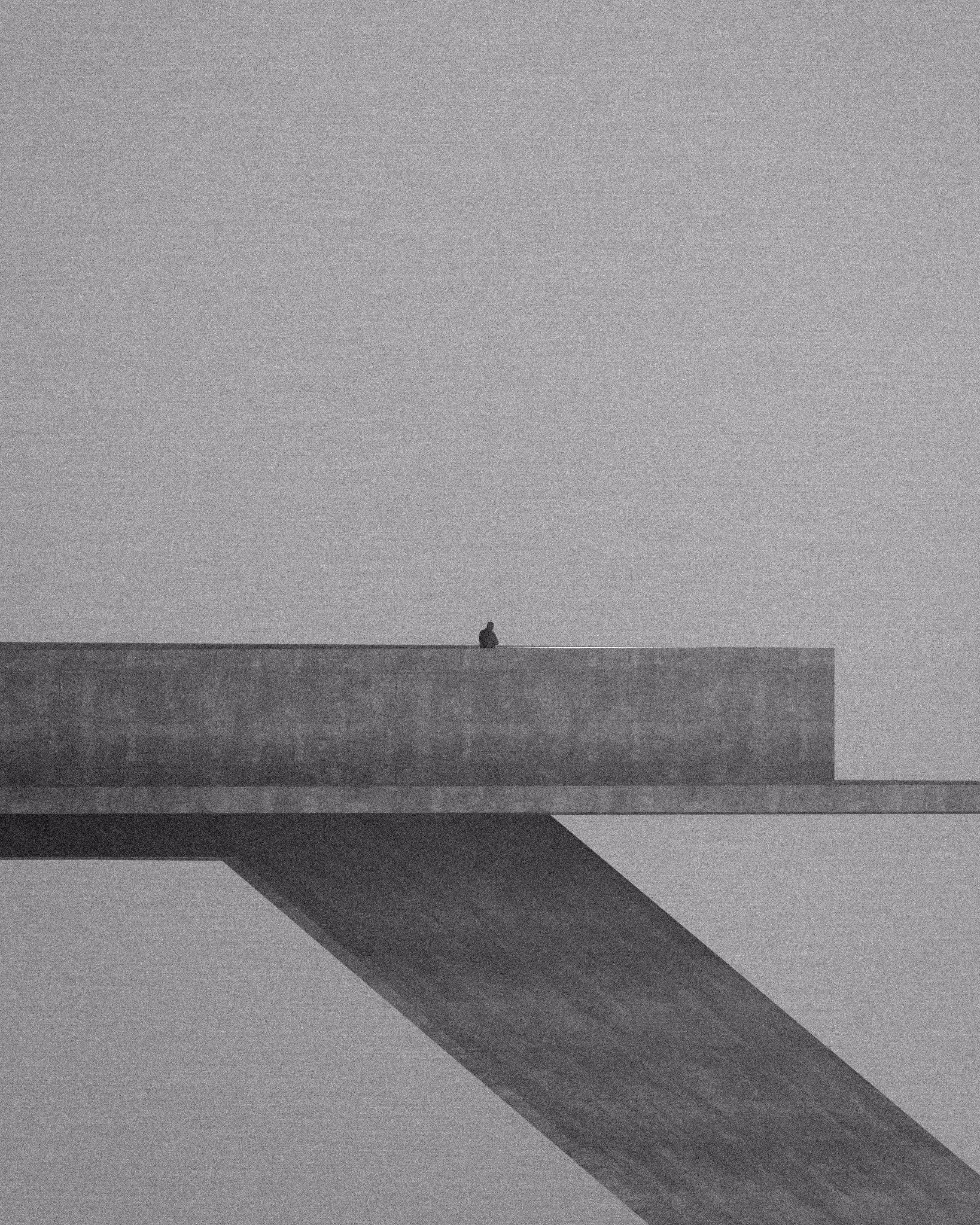 A solitary figure sits on a long concrete beam, casting a monumental diagonal shadow across a pale field.